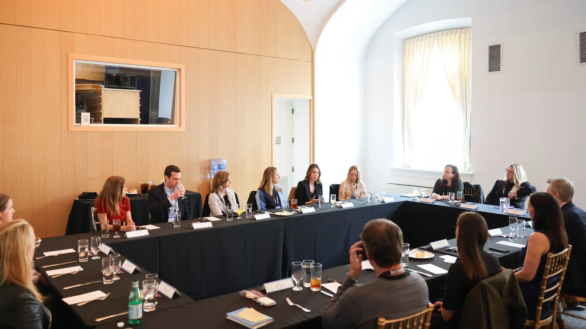 A professional panel discussion in a bright conference room: people seated around a U-shaped table with name cards, glasses, and drinks; wood-paneled wall and a large arched window.