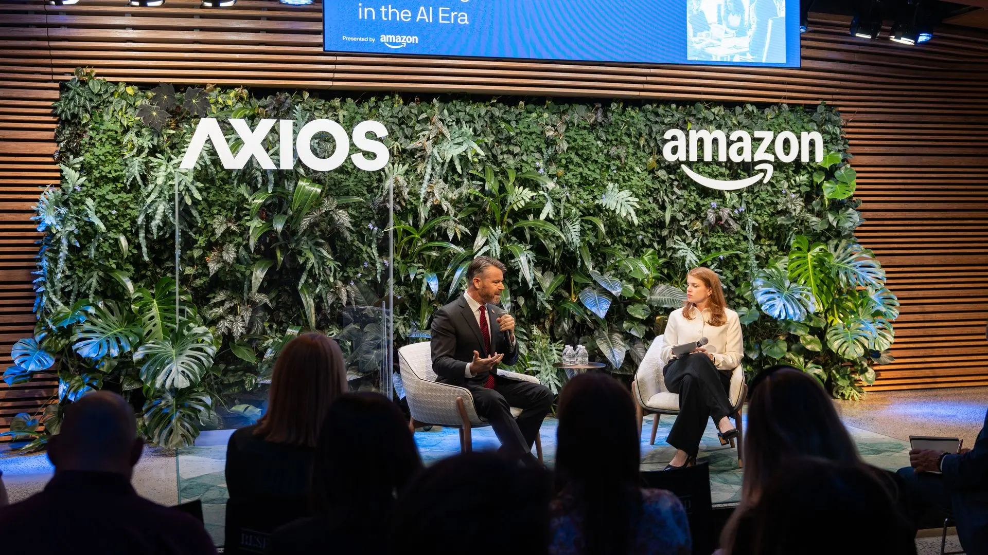 Panel discussion on a stage with a green plant wall and Axios and Amazon logos; a blue screen displays "Future of Work: Succeeding in the AI Era" as two speakers converse.