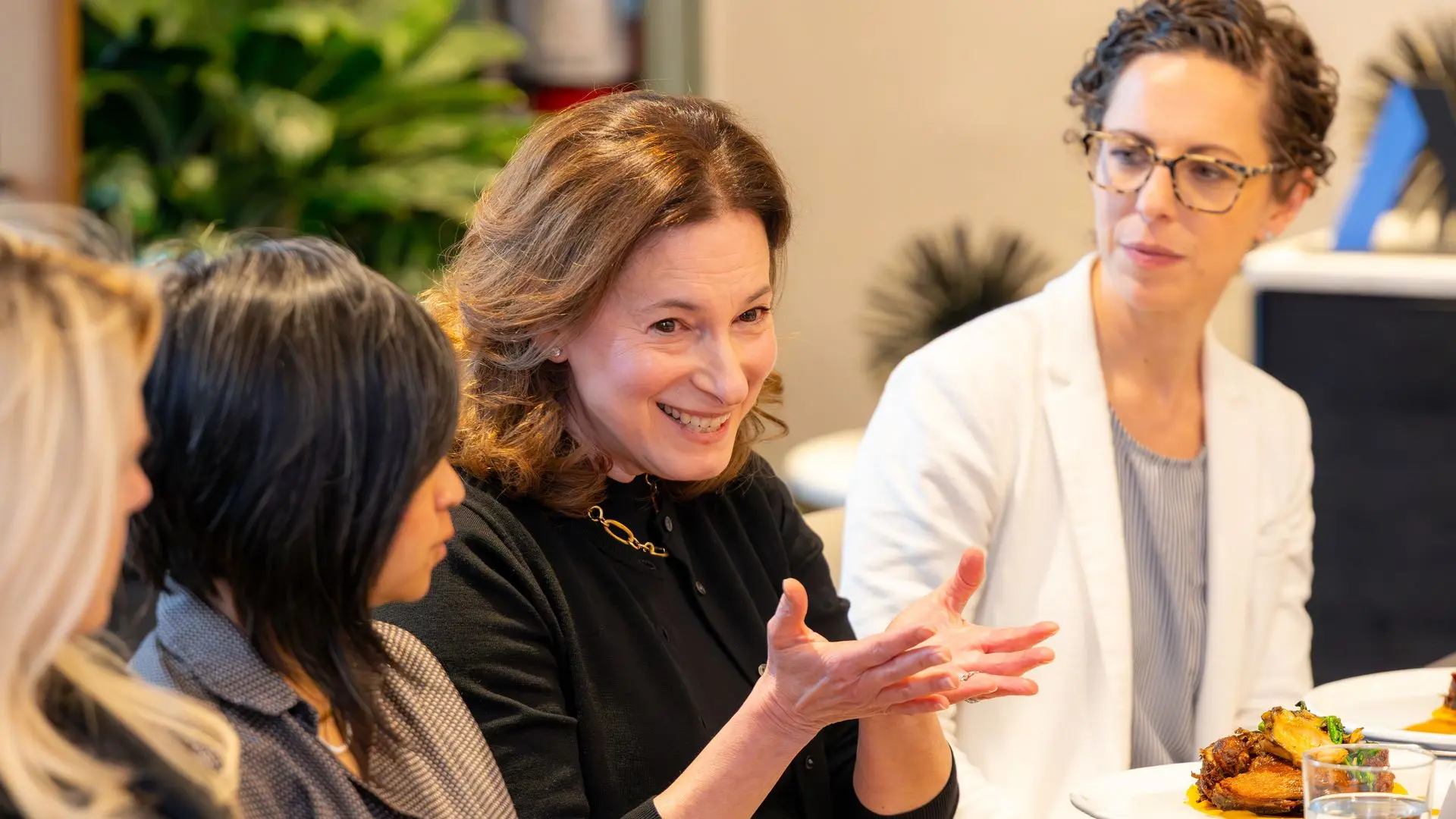 Three women sit at a table in a bright office, talking. Center woman smiles in a black top; right, a woman with glasses and a white blazer. Plates and drinks visible in foreground.