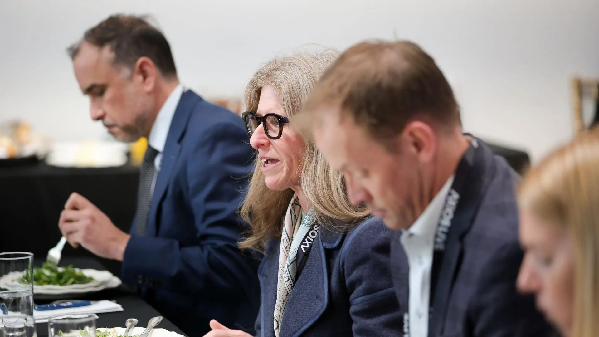 Three adults at a formal luncheon sit at a black-clothed table; a blonde woman with glasses and a scarf speaks, while two men in suits eat salad beside her.