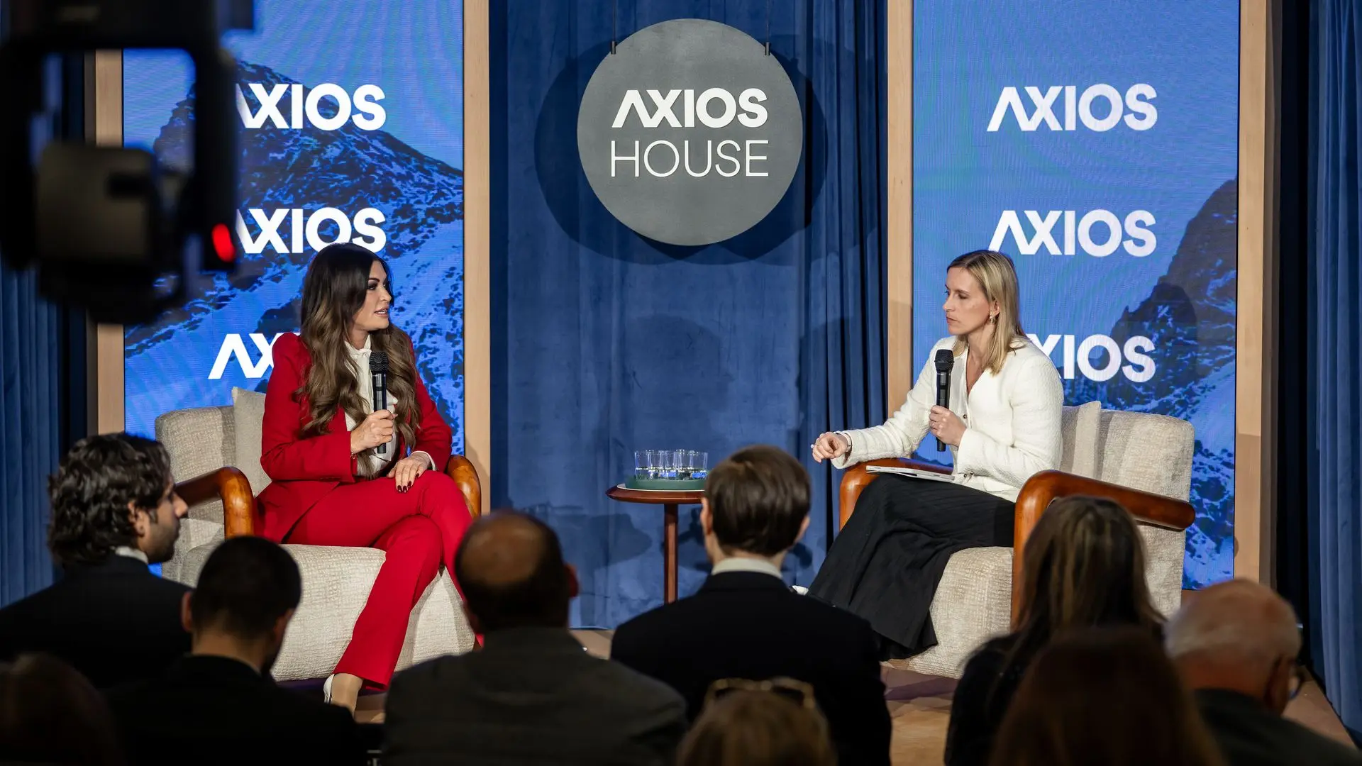 Two women seated on stage at Axios House event, holding microphones; one in a red suit, the other in white top. Audience watches with Axios logos on blue screens behind them.