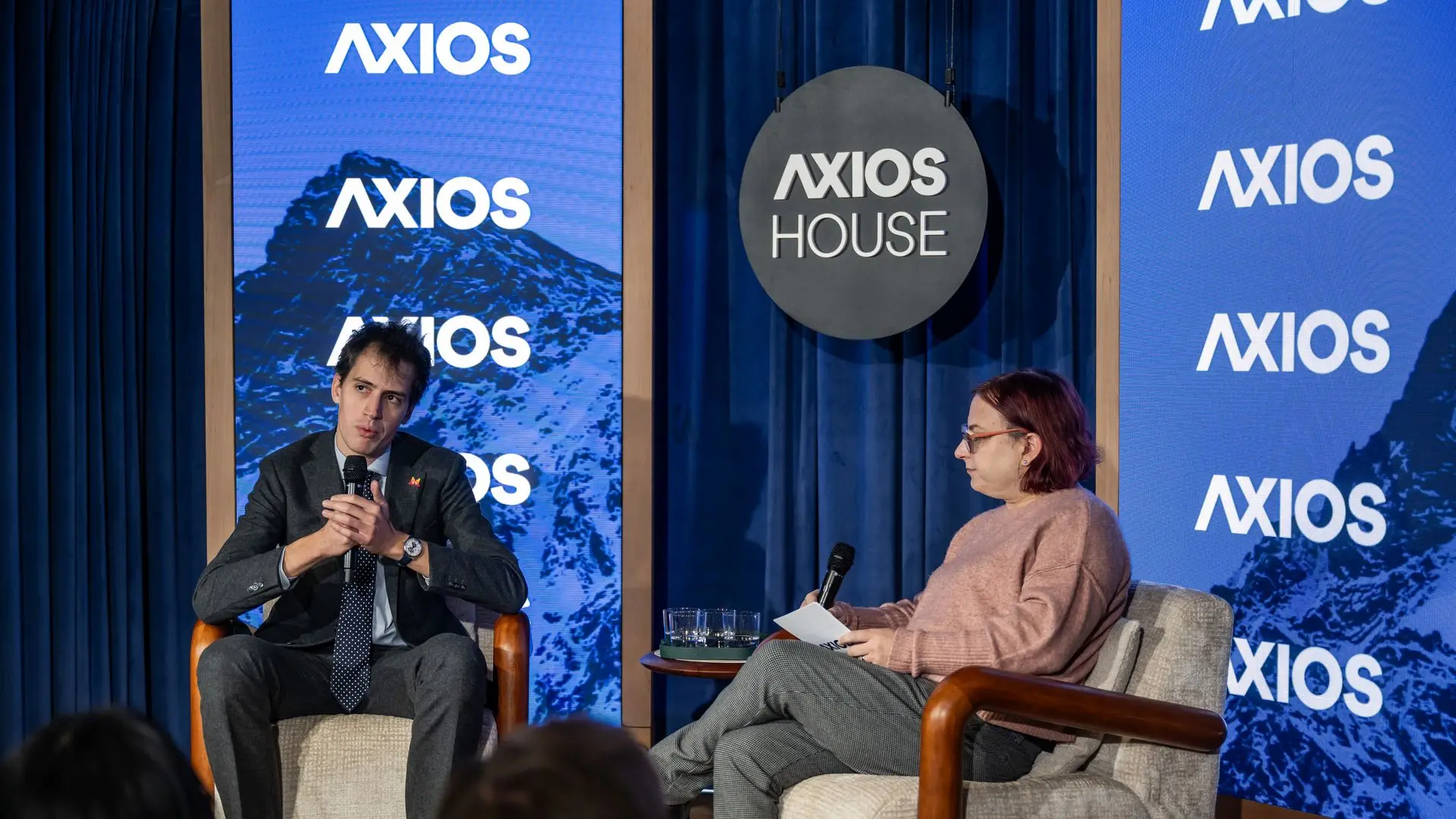 Two people seated on beige chairs on a stage with blue curtains and multiple 'AXIOS' logos on screens behind them. One man in a suit speaks into a microphone, a woman in glasses and a pink sweater holds a microphone and papers.