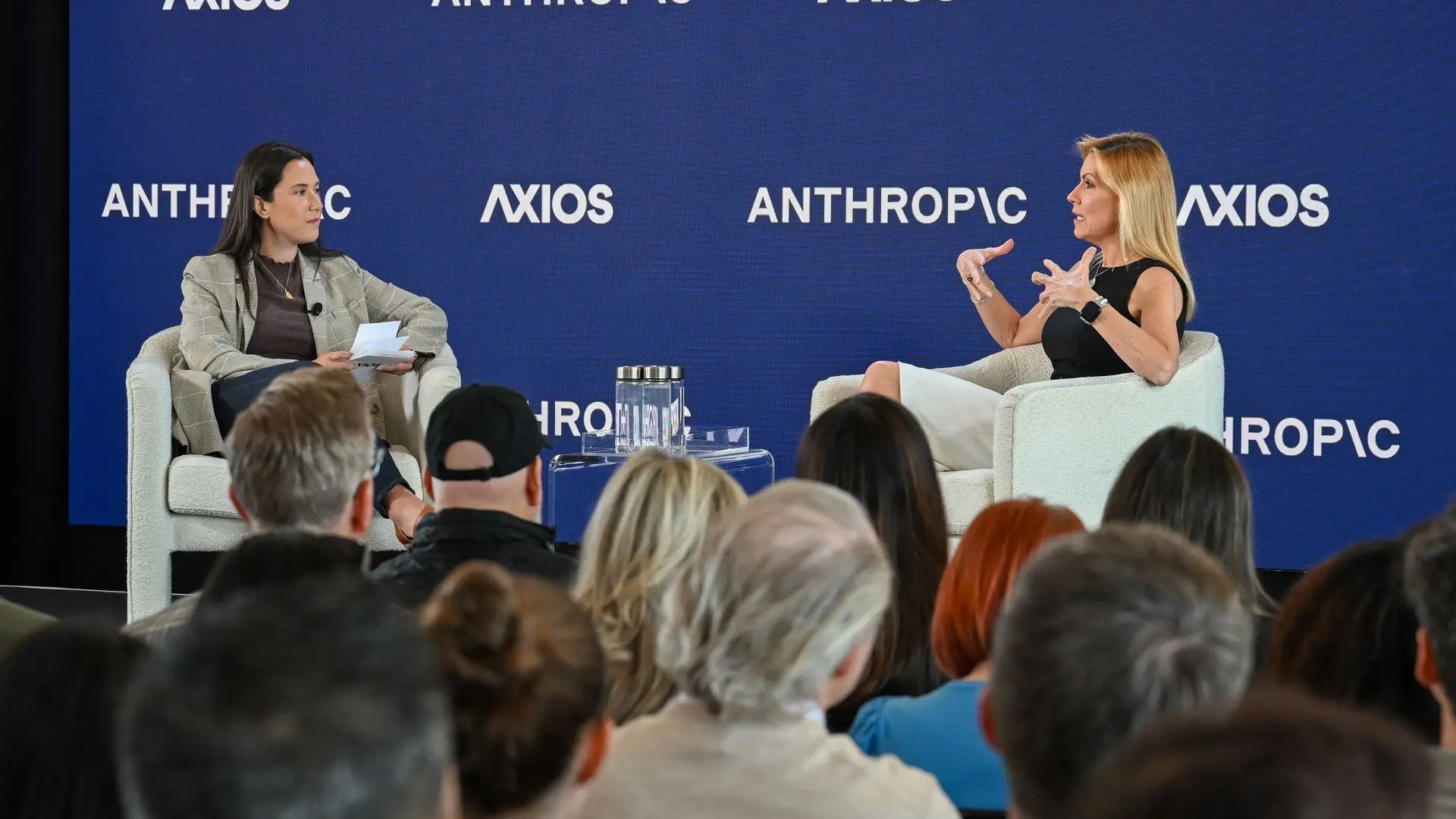 Two women sit on a stage with a blue backdrop displaying AXIOS and ANTHROPIC logos. Left: gray blazer, papers; right: black top and white pants, gesturing as she speaks. Audience in foreground.