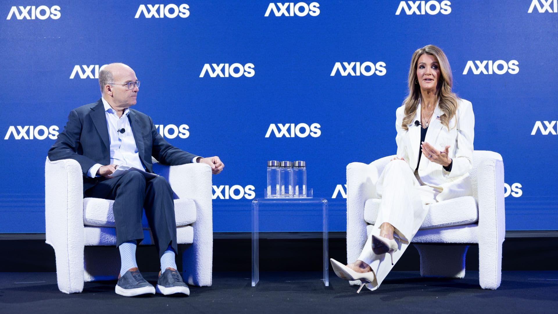 Two panelists sit on white armchairs on a stage with a blue backdrop repeating "AXIOS"; a man in a suit on the left and a woman in a white suit on the right, with a glass table and water bottles between them.